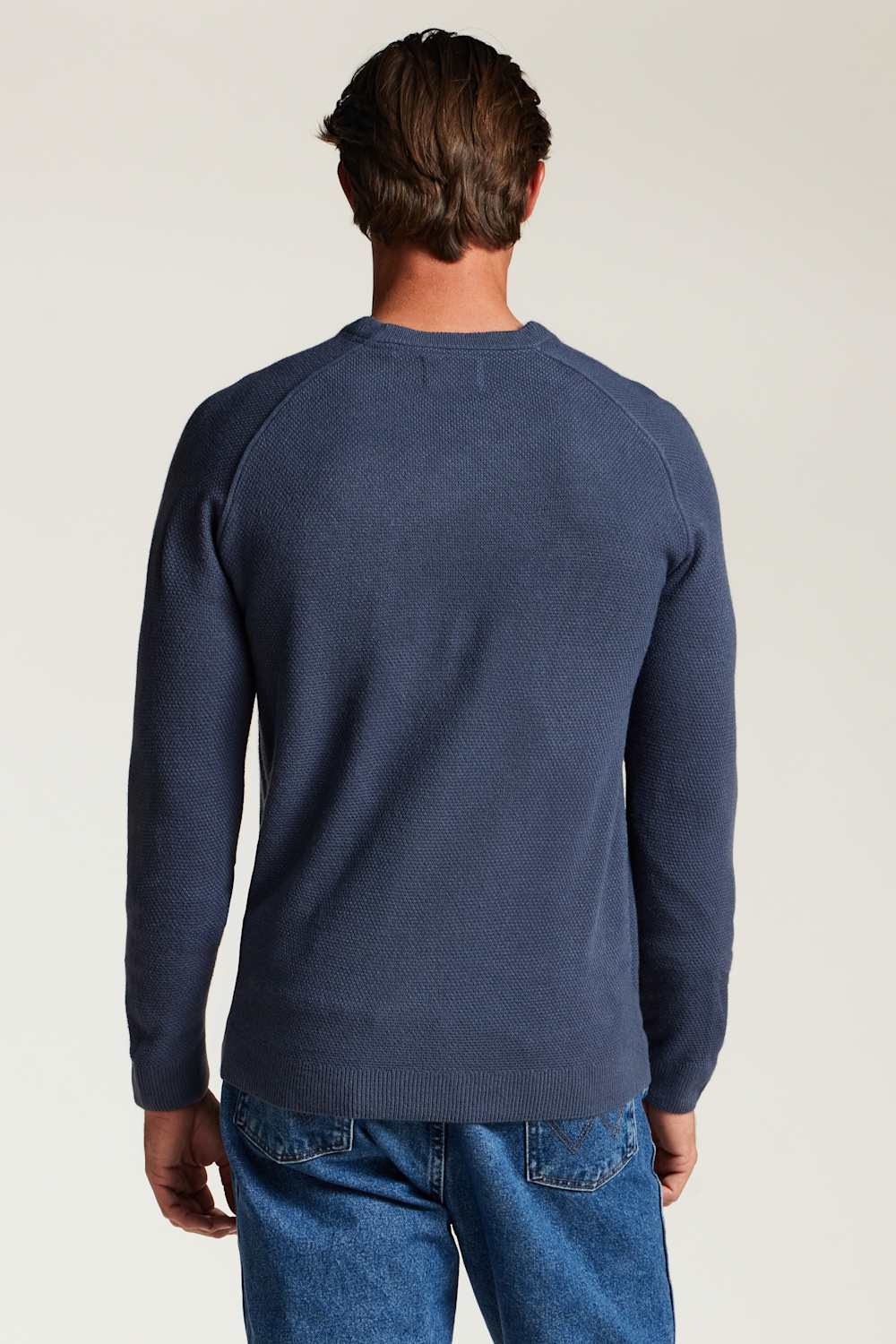 A man viewed from behind wearing a navy blue long-sleeve crewneck sweater and blue jeans against a plain light background.