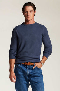 A man wearing a navy blue long-sleeve crewneck sweater, blue jeans, and a brown braided belt, standing against a light beige studio background.