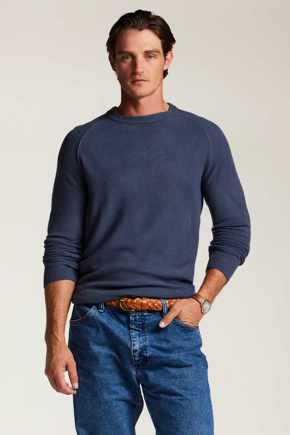 A man wearing a navy blue long-sleeve crewneck sweater, blue jeans, and a brown braided belt, standing against a light beige studio background.