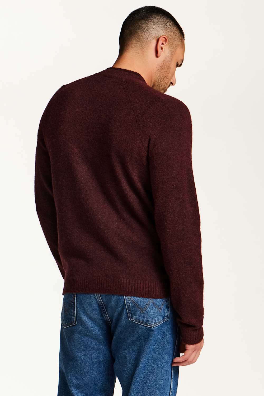 A man viewed from behind wearing a dark burgundy knit sweater and blue jeans against a plain light background.