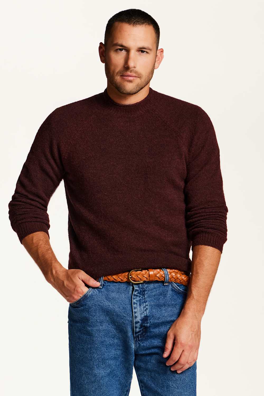 A man wearing a dark burgundy knit crewneck sweater, blue jeans, and a brown braided belt, standing against a plain off-white background.