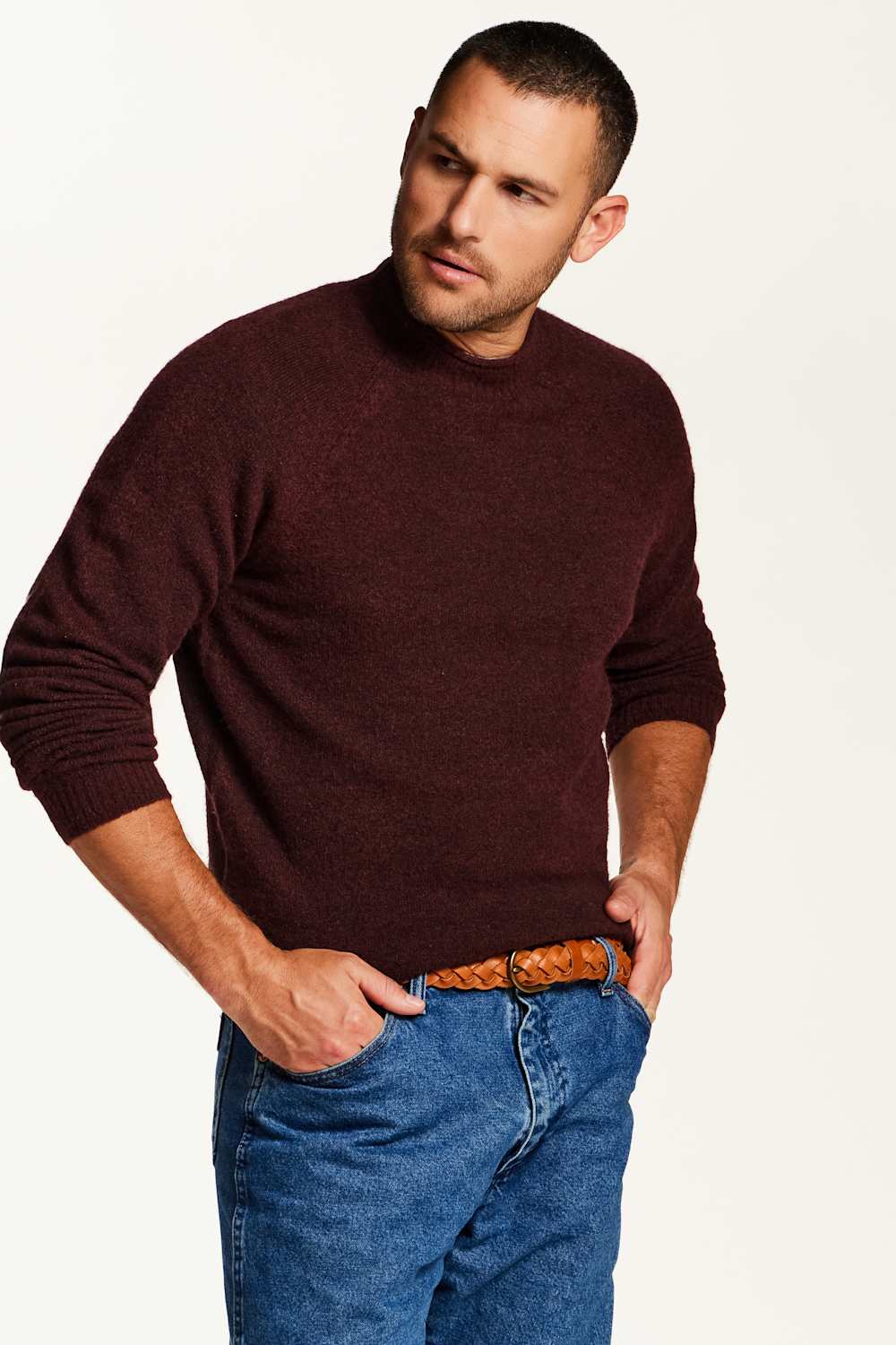 A man wearing a dark burgundy knit sweater and blue jeans with a brown woven belt, standing against a plain white studio background.