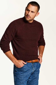 A man wearing a dark burgundy knit sweater and blue jeans with a brown woven belt, standing against a plain white studio background.