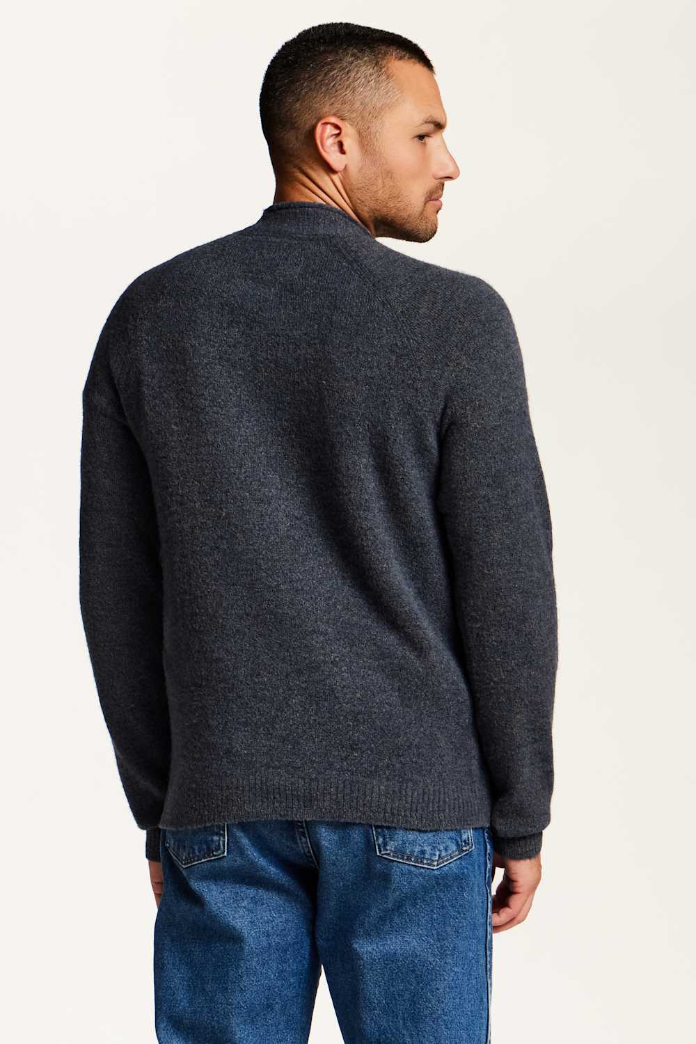 A man wearing a dark charcoal gray knit sweater and blue jeans, shown from the back against a plain off-white background.