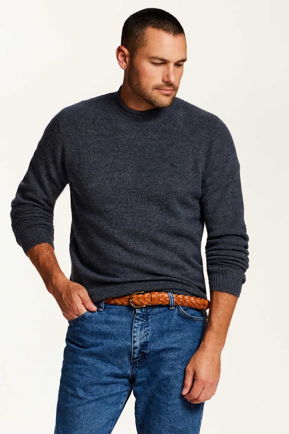A man wearing a dark heather gray crewneck sweater, blue jeans, and a brown woven belt, photographed against a plain off-white studio background.