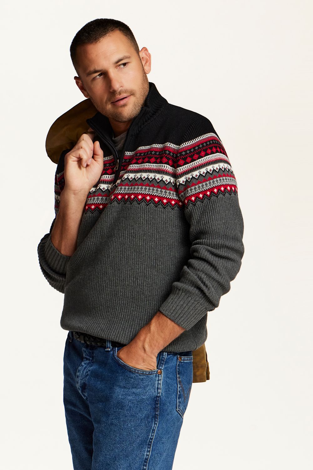 A man wearing a dark gray ribbed knit quarter-zip sweater with a black yoke and a red, white, and black geometric Fair Isle pattern across the chest, paired with blue jeans against a plain off-white background.