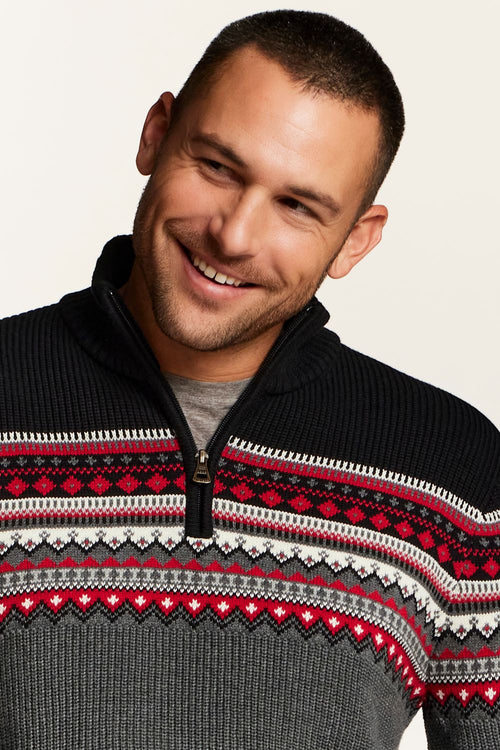 A smiling man wearing a dark gray and black quarter-zip knit sweater with a red, white, and gray Fair Isle pattern across the chest, over a gray t-shirt, against a light beige background.