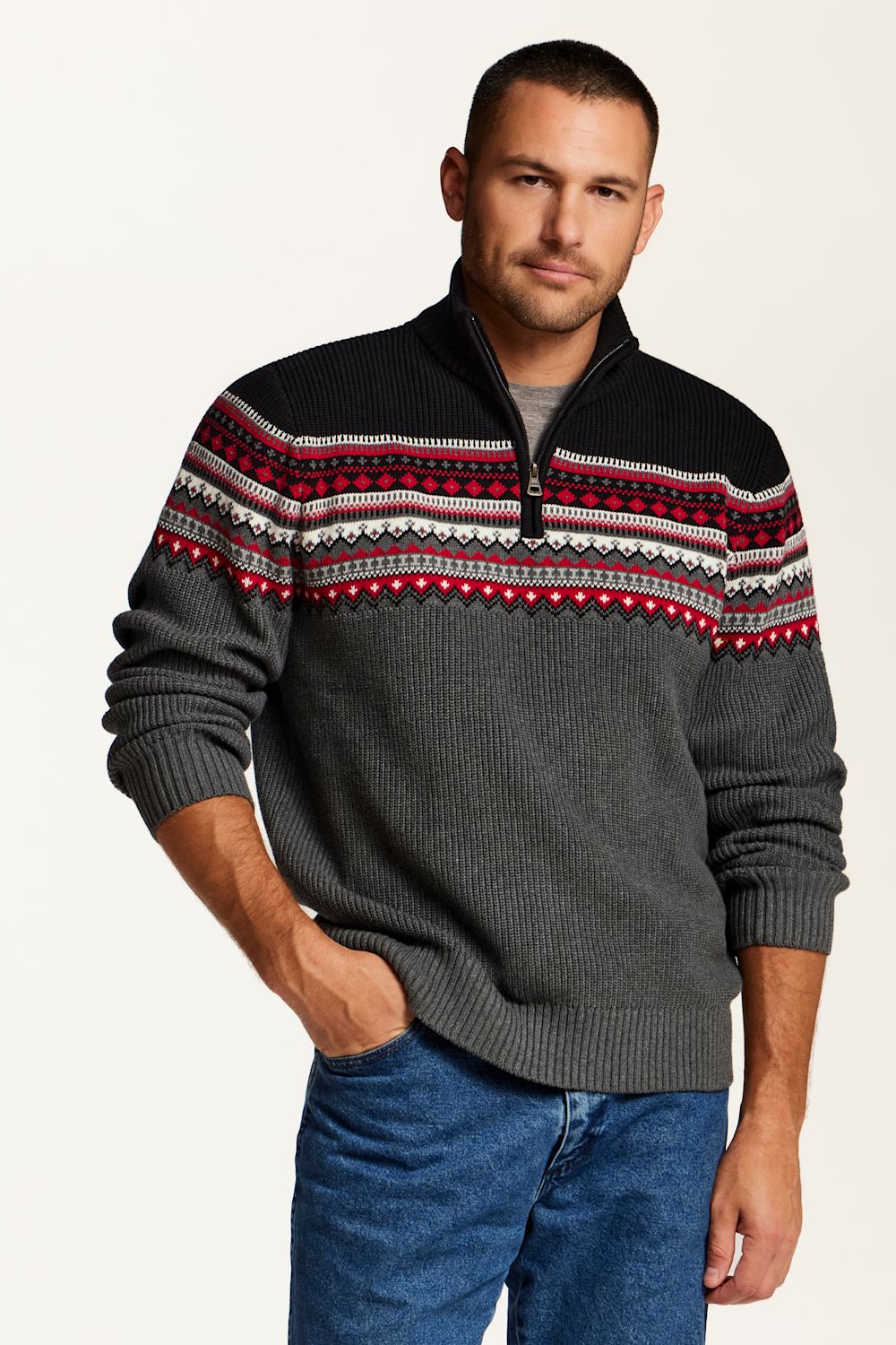 A man wearing a dark gray ribbed knit quarter-zip sweater with a black yoke and a red, white, and gray Fair Isle pattern across the shoulders, paired with blue jeans, against a plain off-white background.