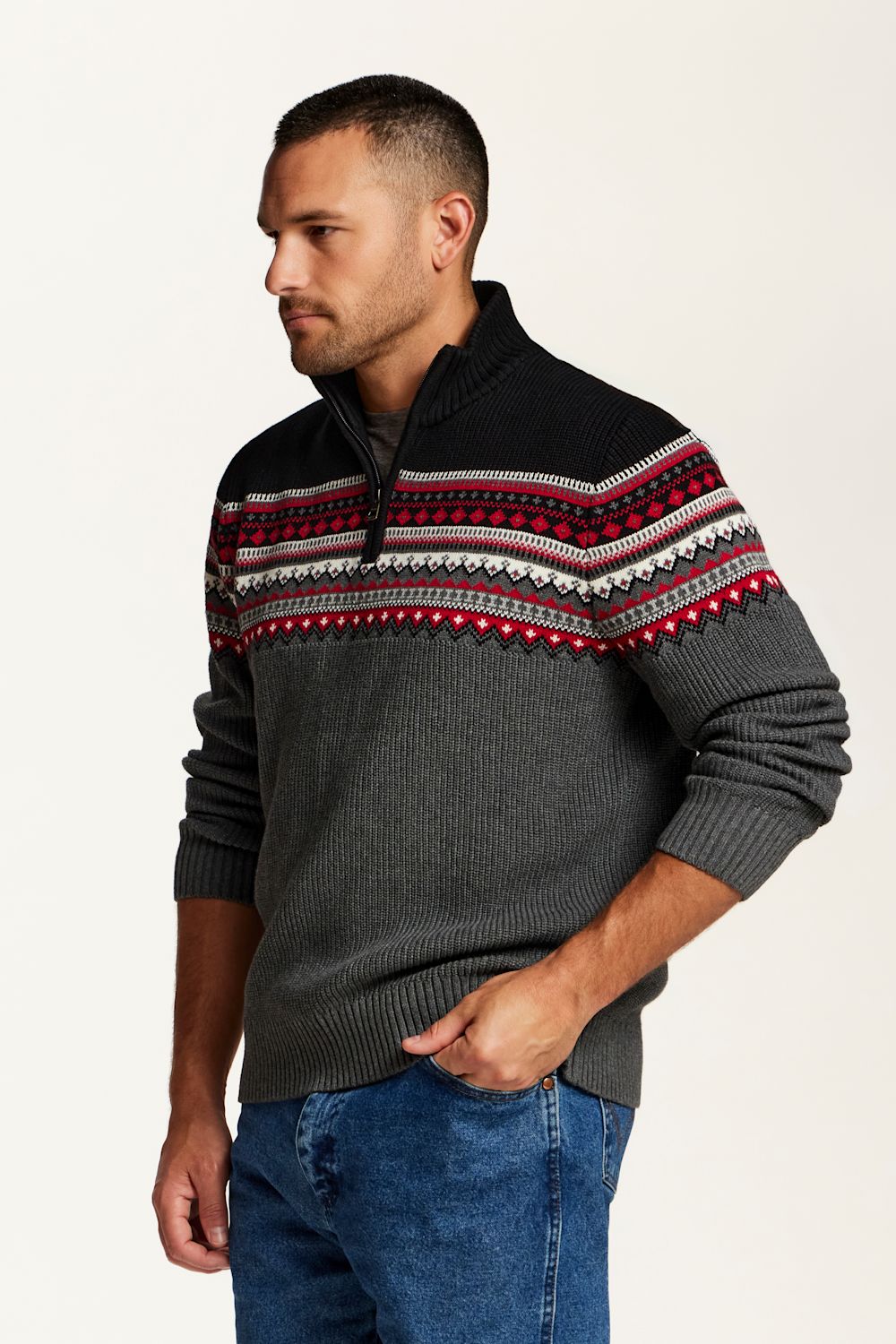 A man wearing a dark gray quarter-zip sweater with a black yoke and a red, white, and gray Fair Isle pattern across the chest, paired with blue jeans, photographed against a plain off-white background.