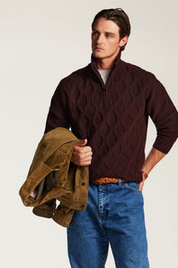 A man wearing a dark burgundy cable-knit quarter-zip sweater and blue jeans, holding a brown suede jacket against a plain light background.