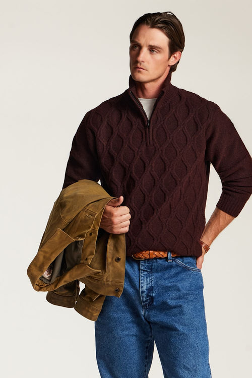 A man wearing a dark burgundy cable-knit quarter-zip sweater and blue jeans, holding a brown suede jacket against a plain light background.