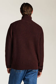 A man viewed from behind wearing a dark burgundy knit sweater and blue jeans, standing against a plain light gray studio background.