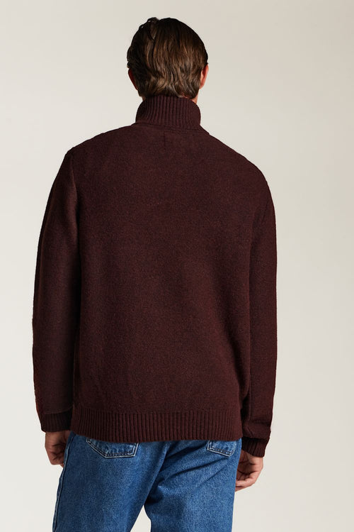 A man viewed from behind wearing a dark burgundy knit sweater and blue jeans, standing against a plain light gray studio background.