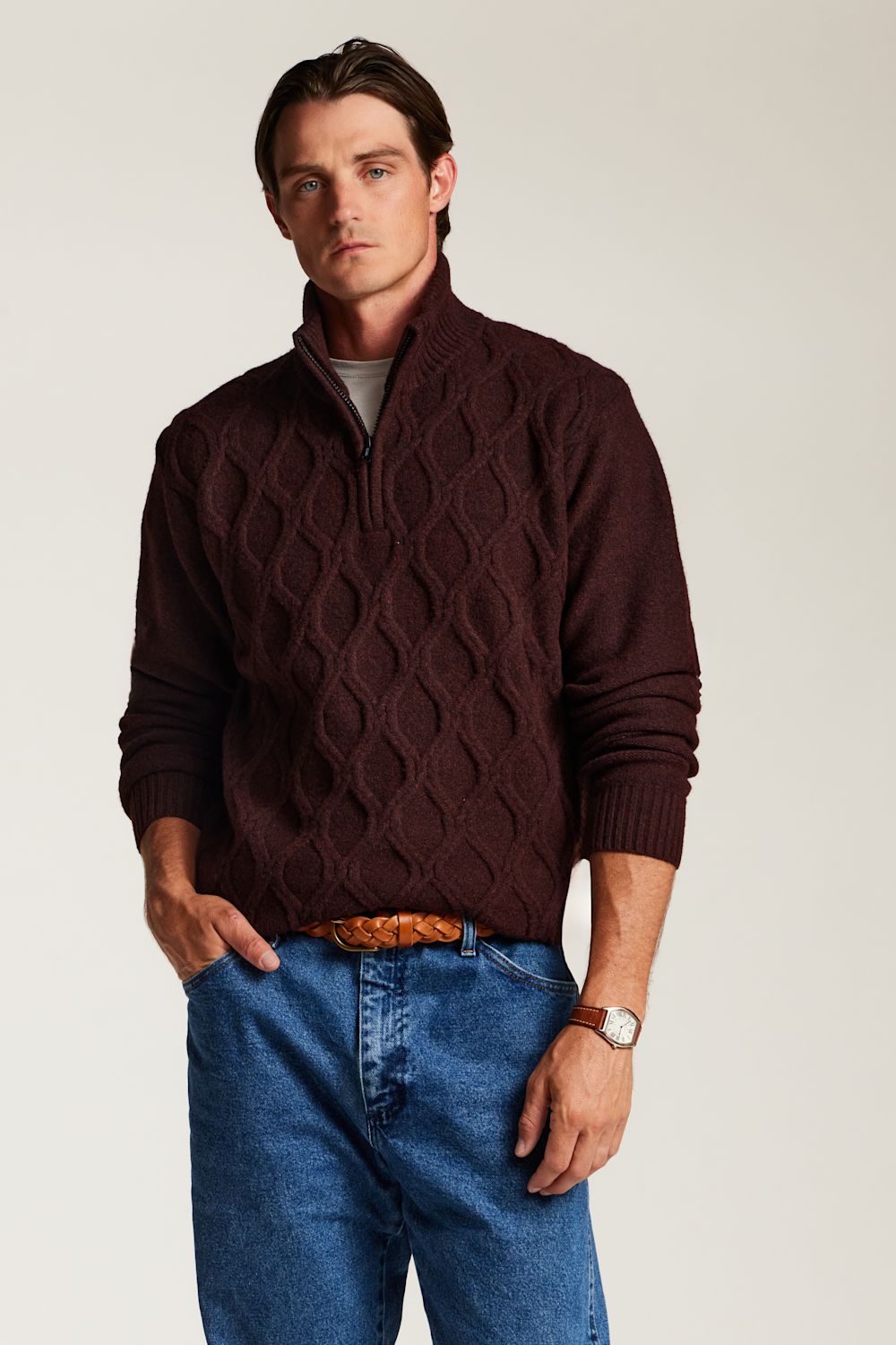 A man wearing a dark maroon cable-knit quarter-zip sweater, blue jeans, and a brown leather belt, standing against a light gray studio background.