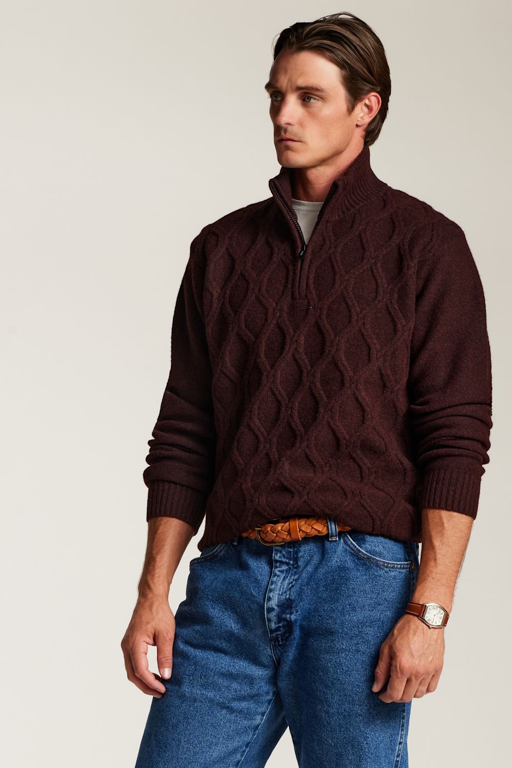 A man wearing a dark maroon cable-knit quarter-zip sweater, blue jeans, and a brown belt, photographed against a plain light gray studio background.