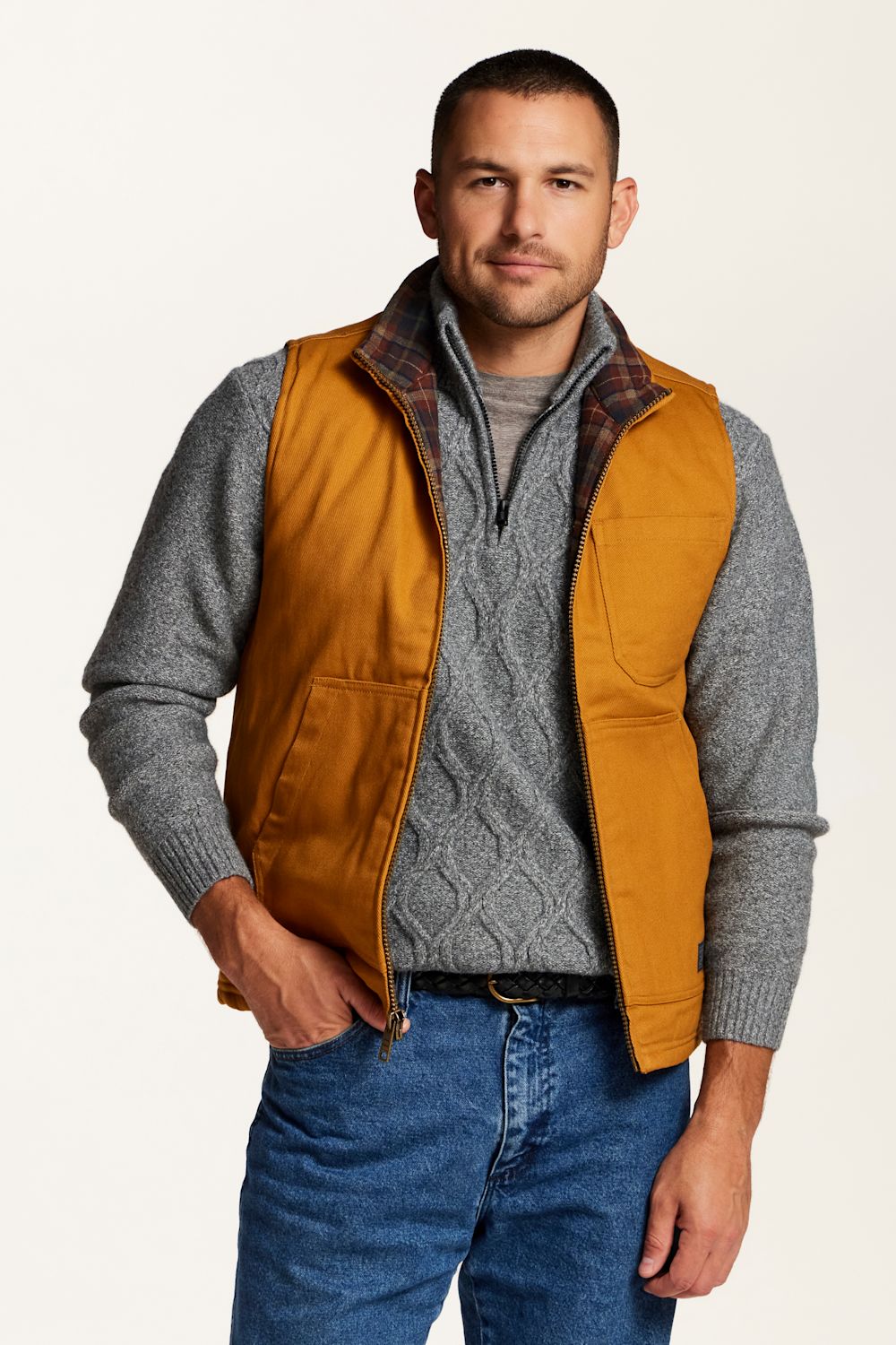 A man wearing a dark yellow zip-up vest over a gray quarter-zip sweater and blue jeans, standing against a plain off-white studio background.