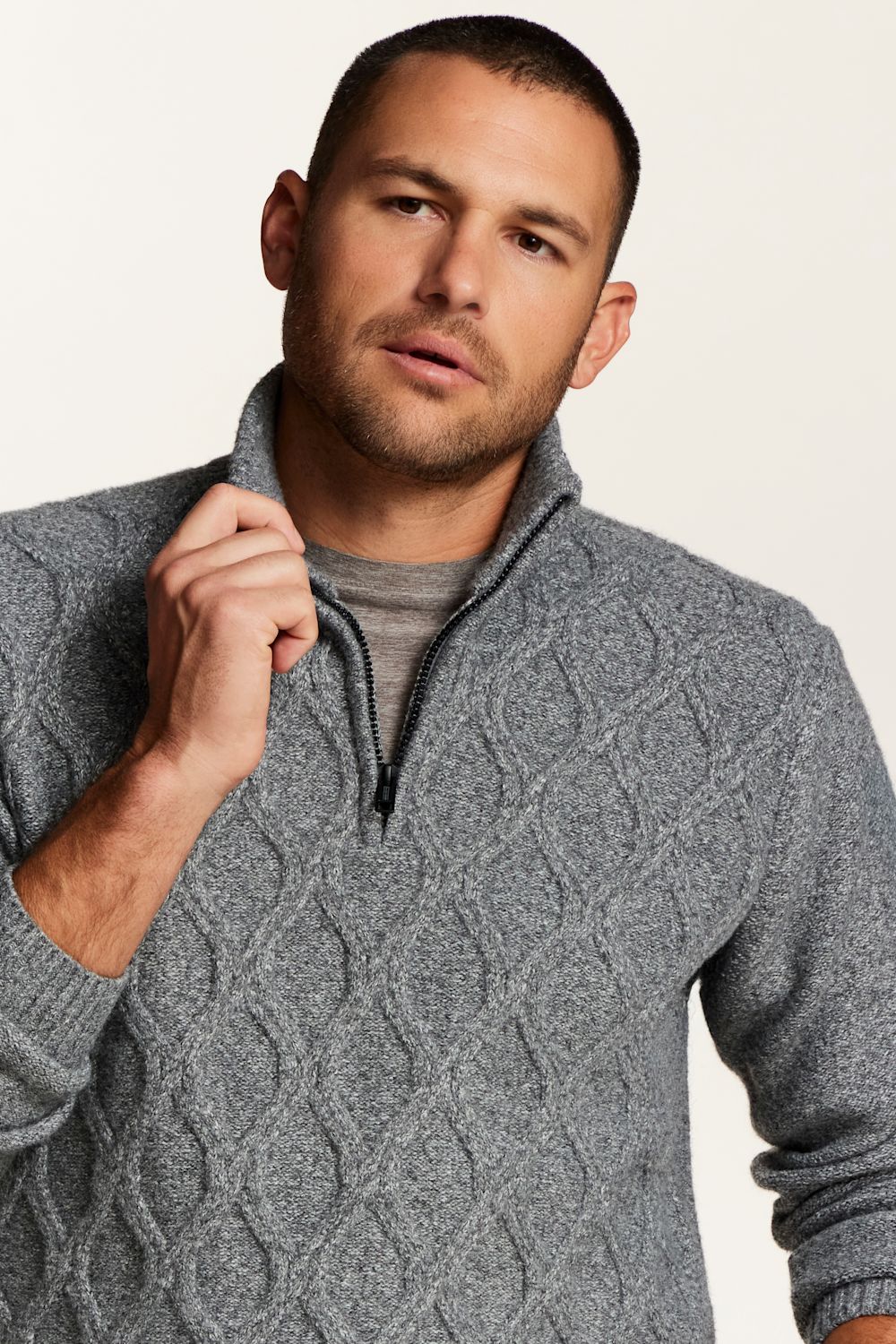 A man wearing a gray cable-knit quarter-zip sweater over a gray t-shirt, photographed against a clean off-white background.