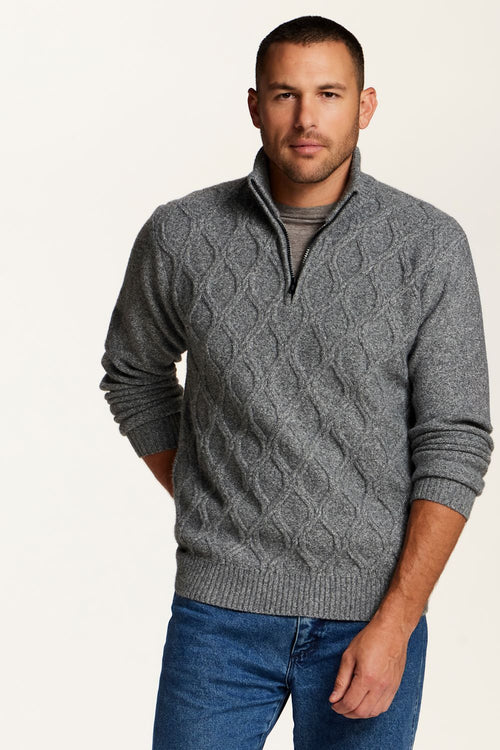 A man wearing a gray cable-knit quarter-zip sweater over a dark gray t-shirt and blue jeans, standing against a plain off-white background.