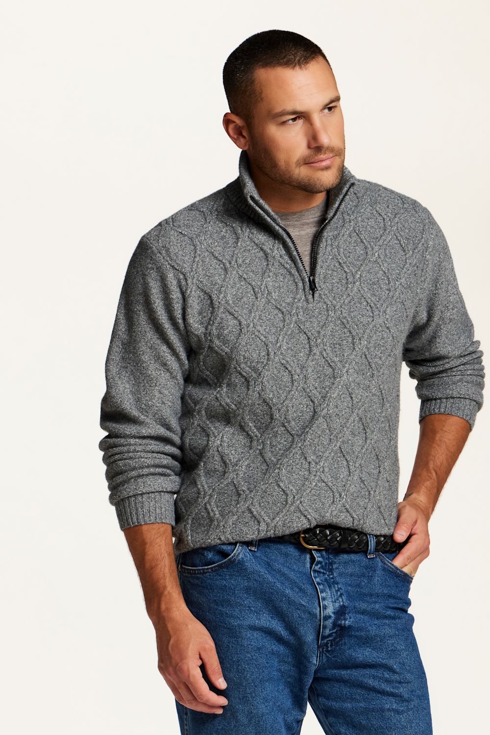 A man wearing a gray cable-knit quarter-zip sweater over a gray t-shirt and blue jeans, standing against a plain off-white background.