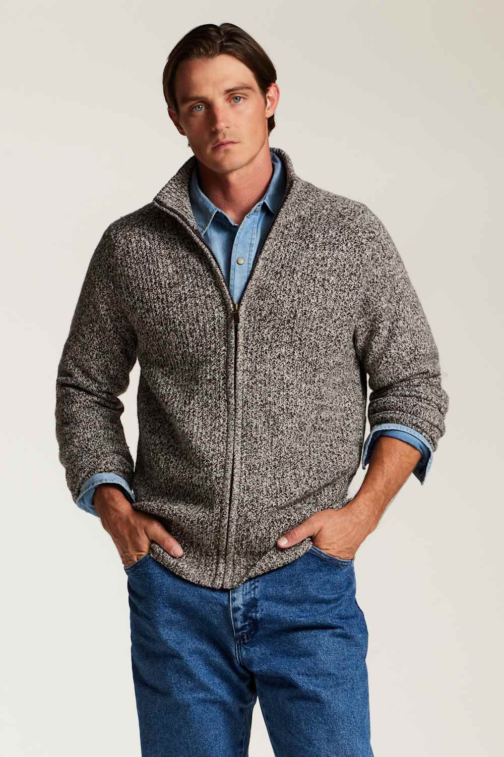 A man wearing a textured gray zip-up sweater over a light blue button-down shirt and blue jeans, standing against a plain light gray studio background.