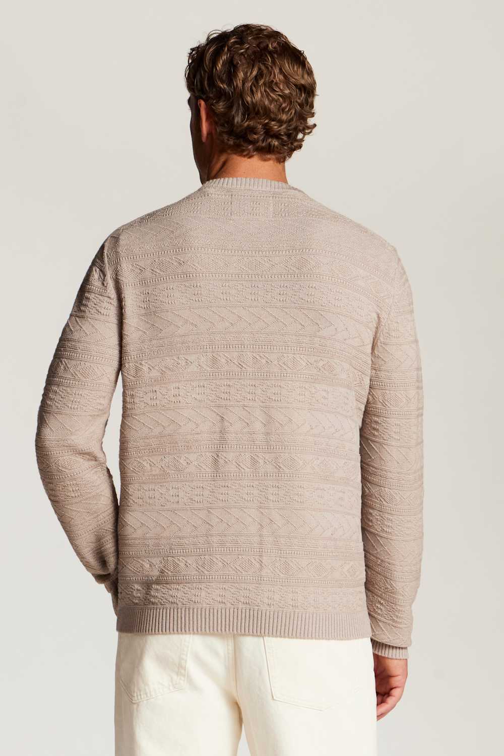 A man viewed from behind wearing a light beige textured knit sweater and off-white trousers, set against a plain light gray studio background.