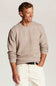 A man wearing a light beige textured knit crewneck sweater, cream-colored trousers, and a black belt, standing against a neutral light gray background.