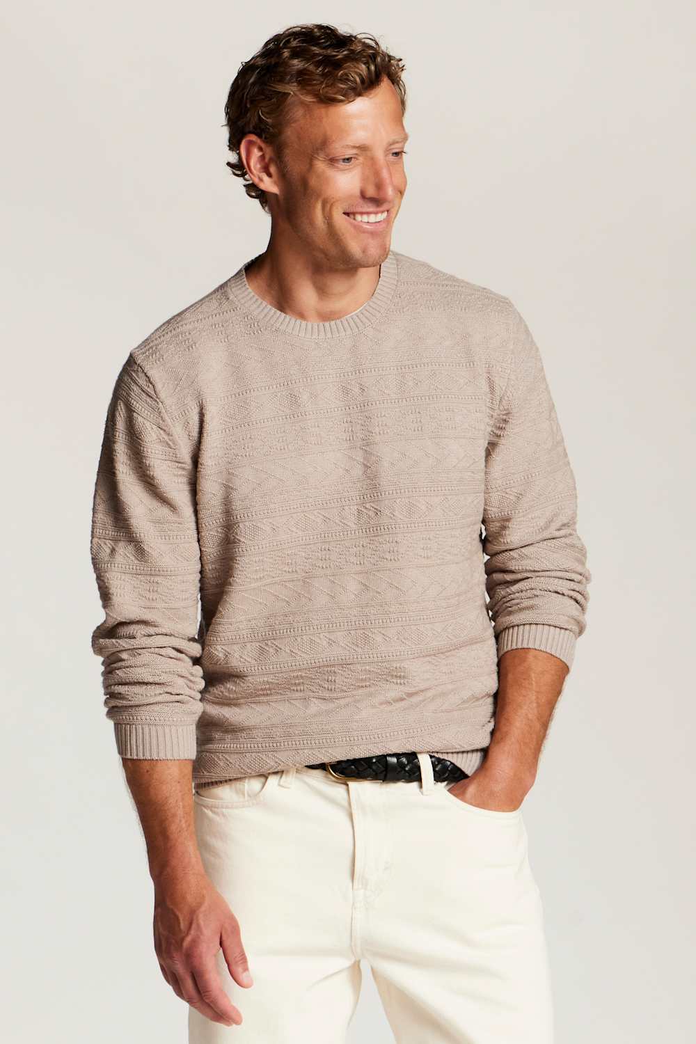 A man wearing a light beige textured knit crewneck sweater, cream-colored trousers, and a black belt, standing against a neutral light gray background.