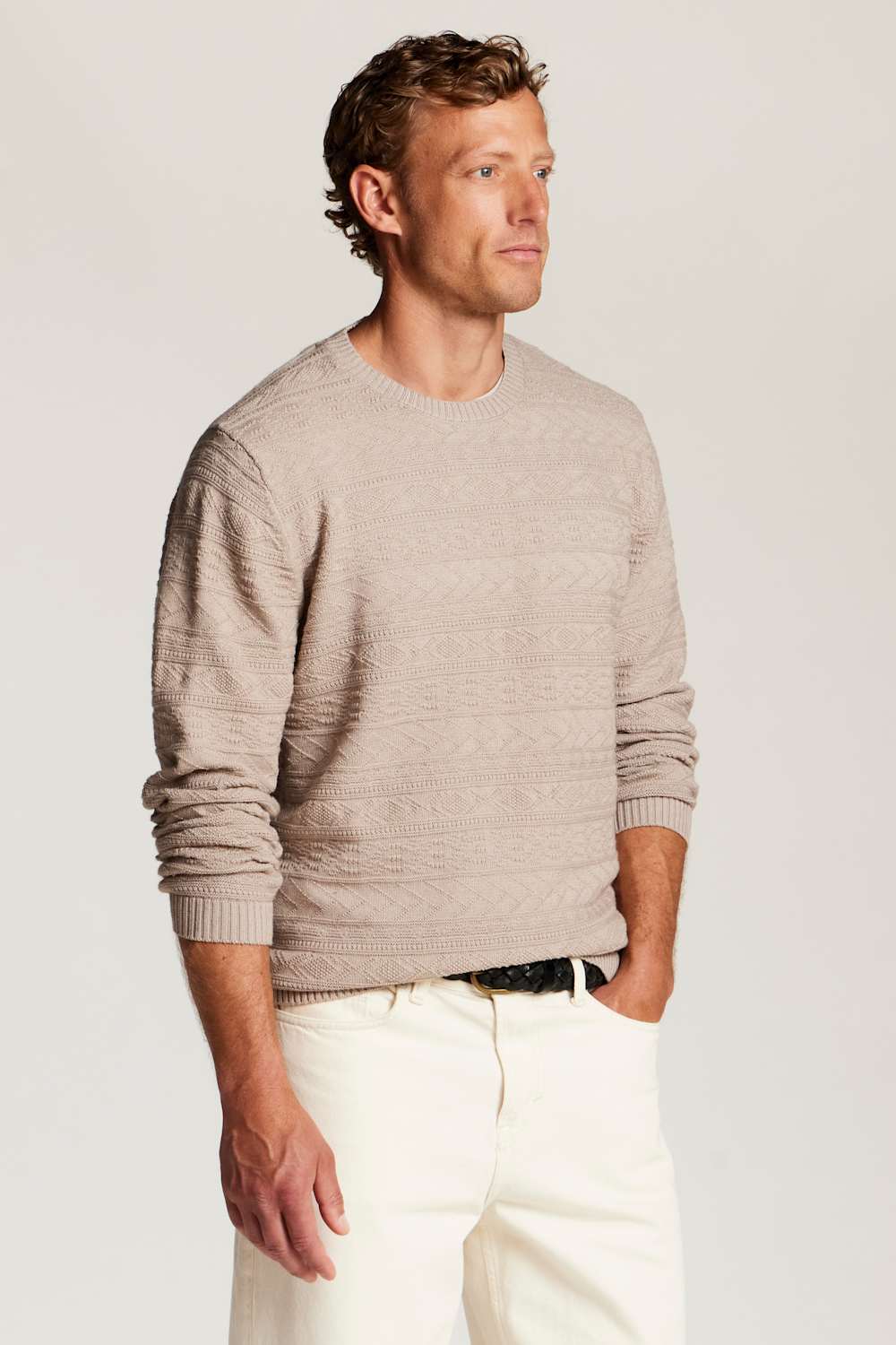 A man wearing a light beige textured knit crewneck sweater and white trousers, standing against a plain light gray studio background.