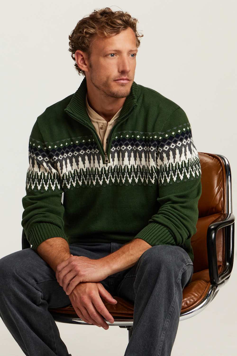 A man wearing a dark green quarter-zip sweater with a white and navy Fair Isle pattern across the chest, sitting on a brown leather chair against a light gray background.