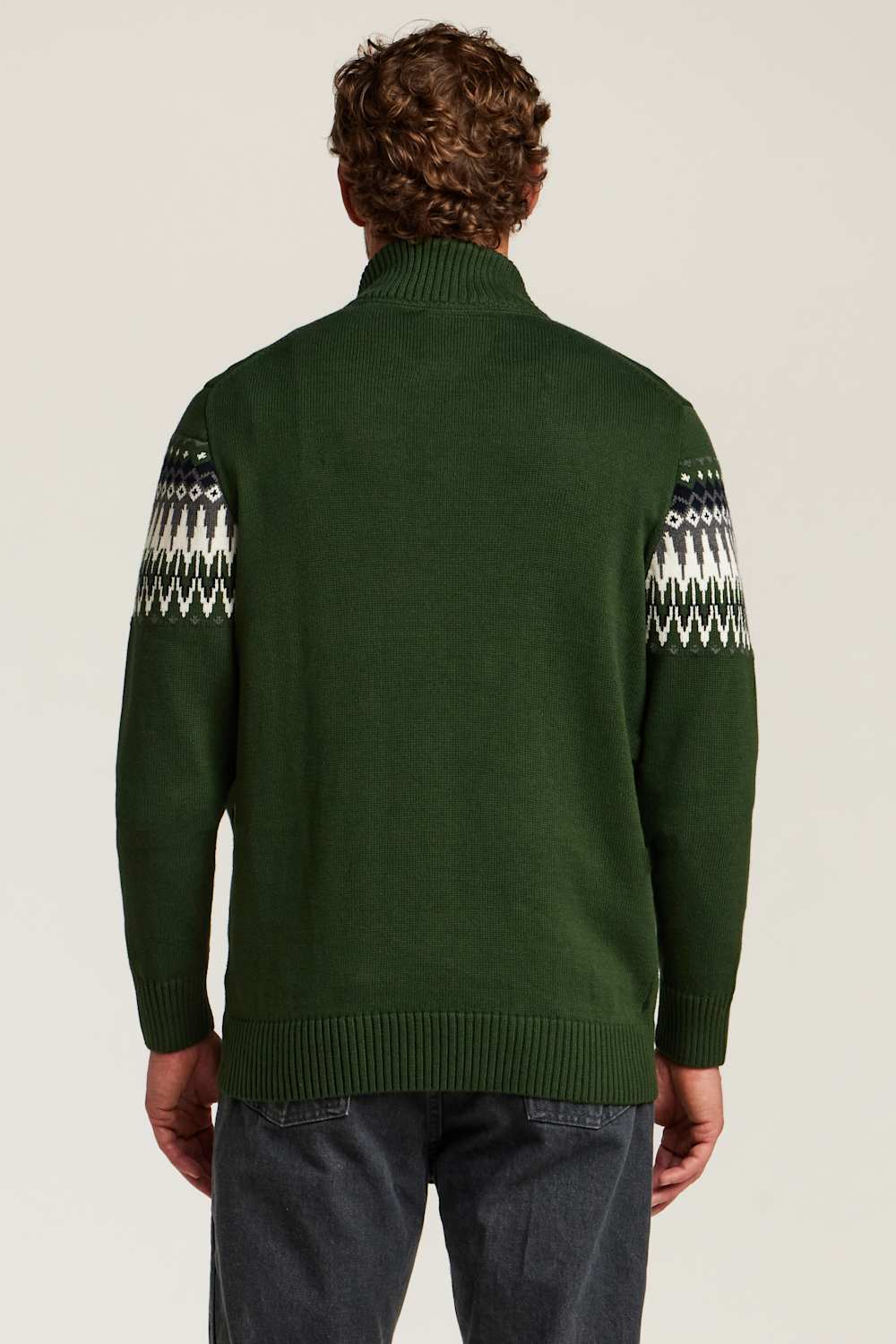 A man viewed from behind wearing a dark green knit sweater with white and gray geometric Fair Isle patterns on the upper sleeves, paired with dark gray jeans, against a light beige studio background.