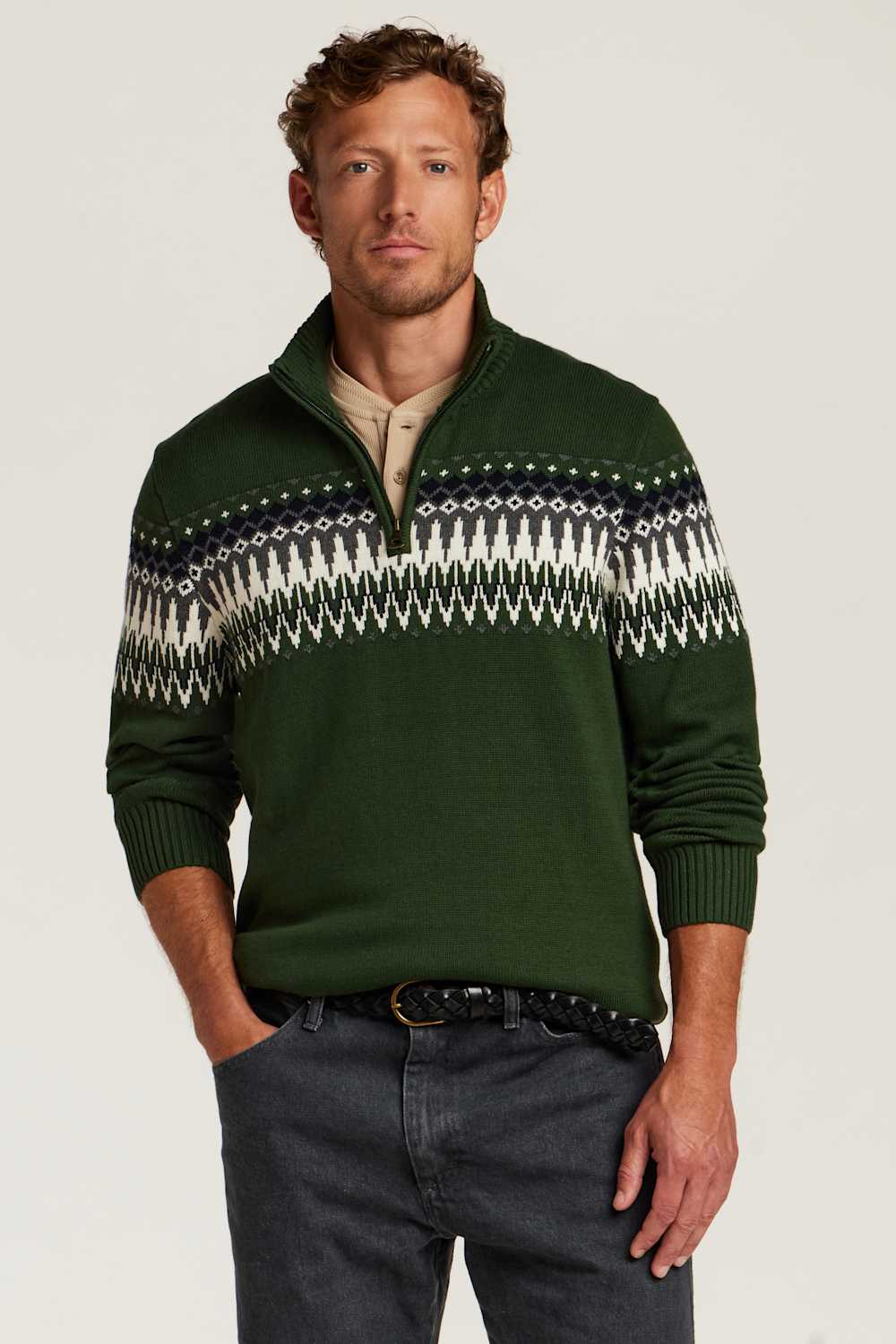 A man wearing a dark green quarter-zip sweater with a white and navy Fair Isle pattern across the chest, paired with dark gray jeans and a black belt, against a light gray studio background.