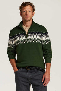 A man wearing a dark green quarter-zip sweater with a white and navy Fair Isle pattern across the chest, paired with dark gray jeans and a black belt, against a light gray studio background.