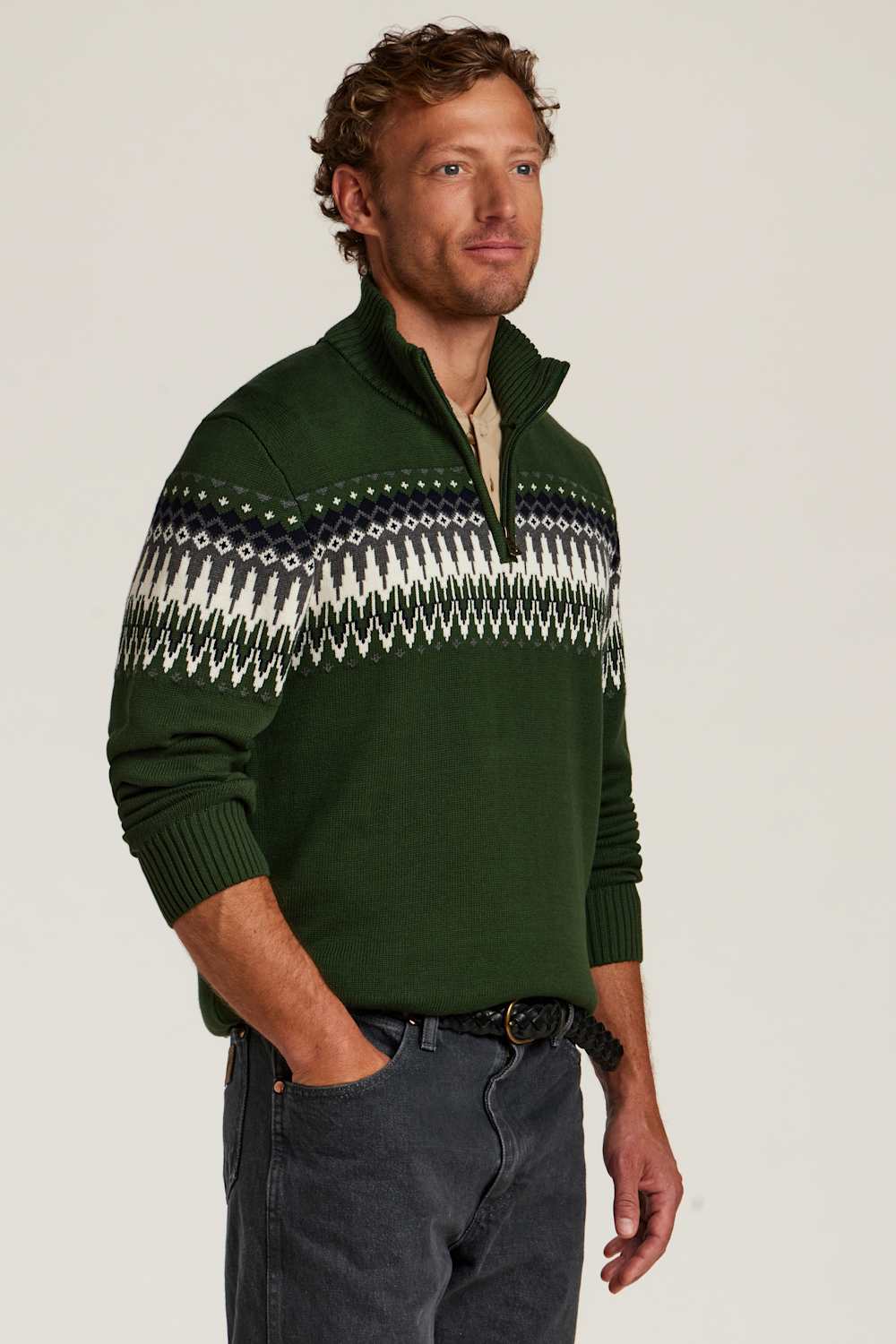 A man wearing a dark green quarter-zip sweater with a white and navy Fair Isle pattern across the chest, paired with dark gray jeans, against a plain light gray studio background.