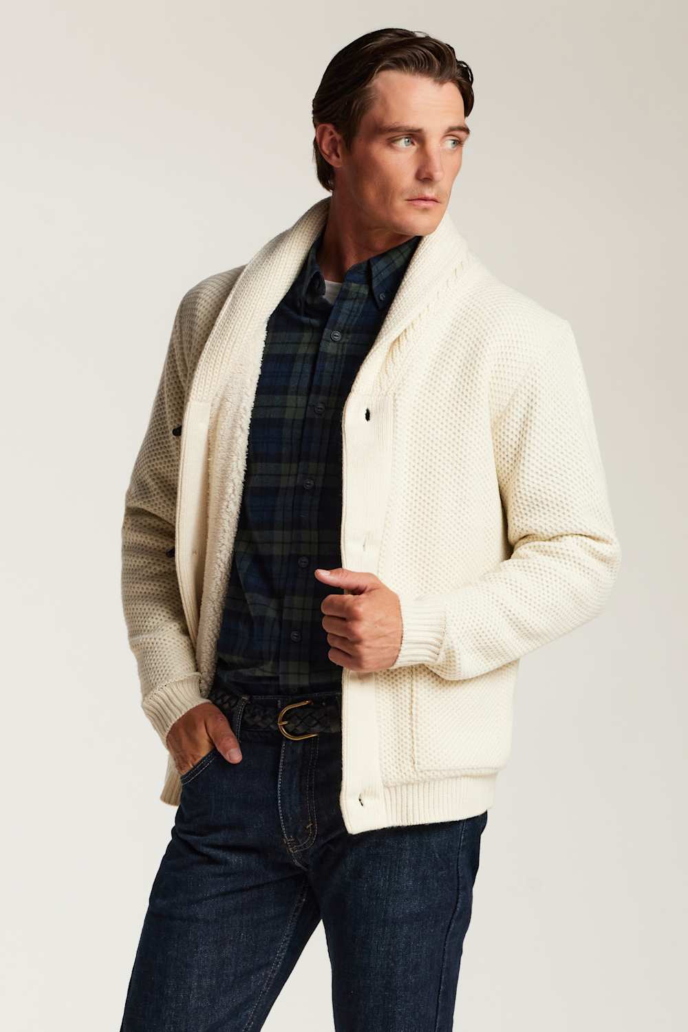 A man wearing a cream-colored textured knit cardigan over a navy and green plaid shirt, paired with dark blue jeans, photographed against a light beige studio background.
