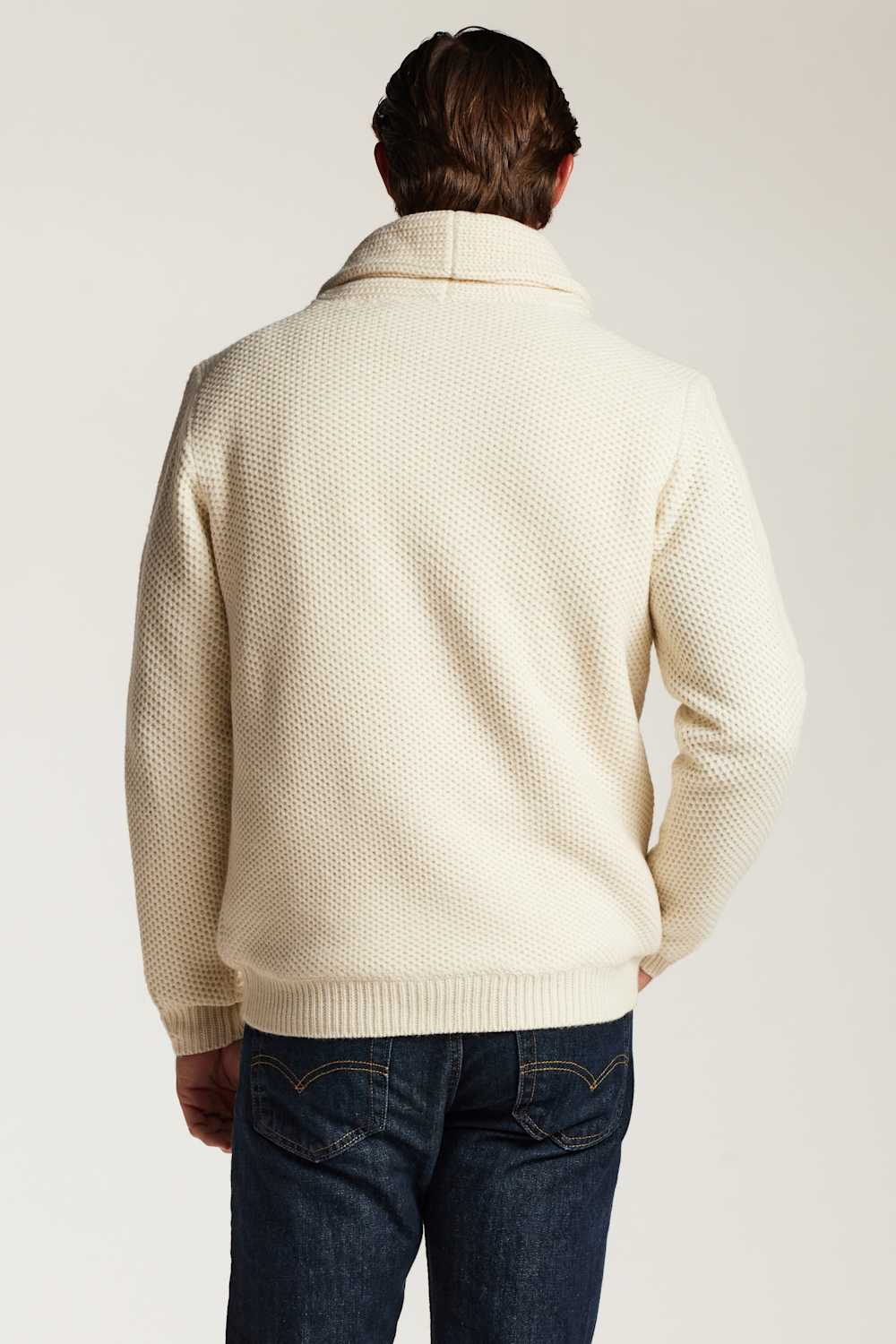 A man viewed from behind wearing a cream-colored textured knit sweater and dark blue jeans against a plain light background.