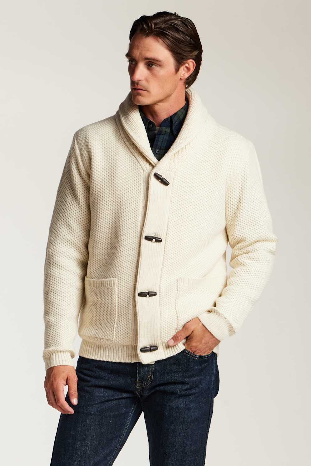 A man wearing a cream-colored textured knit cardigan with toggle closures and a shawl collar, paired with dark blue jeans and a dark plaid shirt, against a light gray studio background.