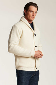 A man wearing a cream-colored textured knit cardigan with a shawl collar, dark buttons, and a ribbed hem, paired with dark blue jeans, photographed against a light gray studio background.