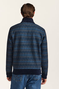 A man viewed from behind wearing a navy blue knit sweater with a blue and white geometric Fair Isle pattern, paired with blue jeans against a plain off-white background.