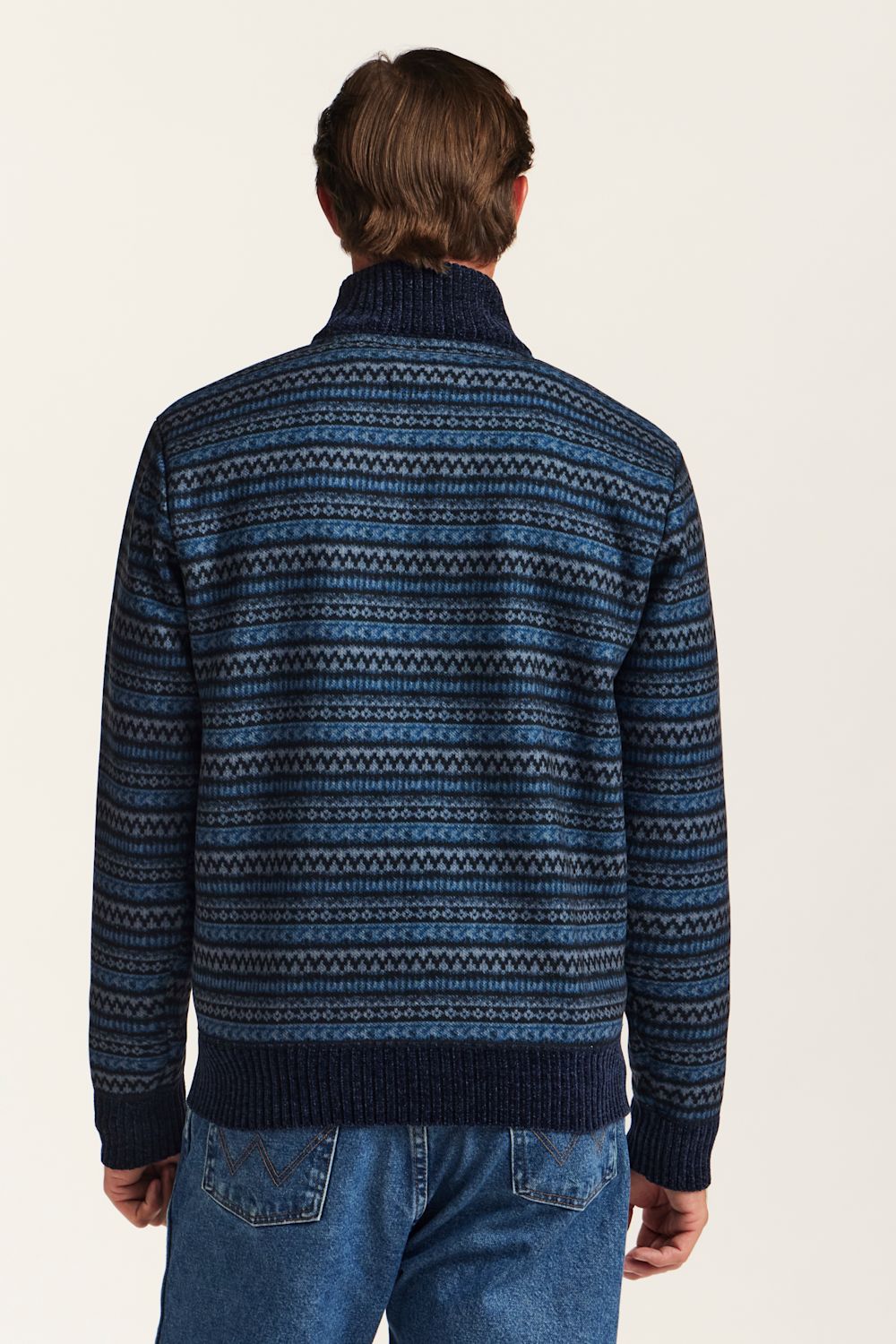 A man viewed from behind wearing a navy blue knit sweater with a blue and white geometric Fair Isle pattern, paired with blue jeans against a plain off-white background.