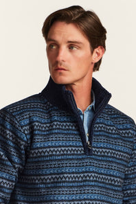 A man wearing a blue and navy patterned quarter-zip sweater over a light blue collared shirt, photographed against a plain light background.
