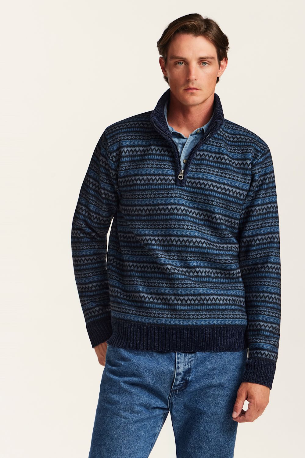 A man wearing a navy blue and blue patterned quarter-zip sweater over a light blue collared shirt and blue jeans, standing against a plain off-white background.