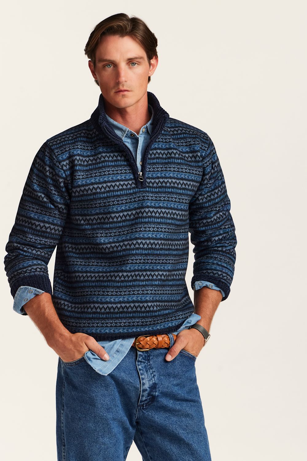 A man wearing a blue and navy patterned quarter-zip sweater over a light blue shirt and blue jeans, standing against a plain off-white background.