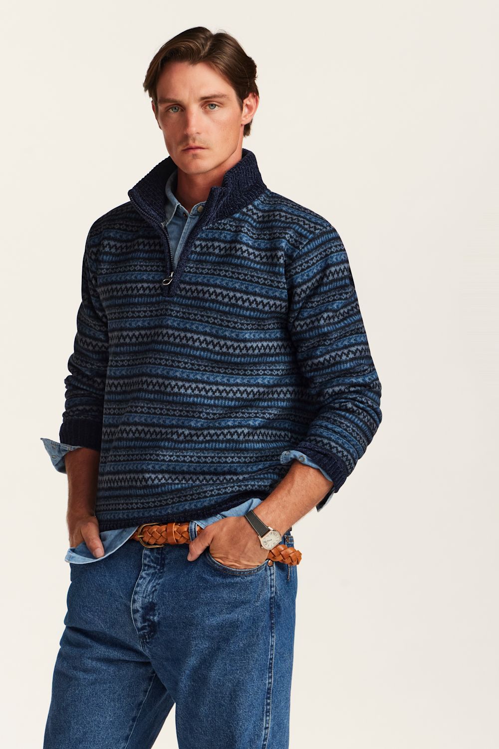 A man wearing a navy blue patterned quarter-zip sweater over a light blue collared shirt, paired with blue jeans and a brown woven belt, photographed against a plain off-white background.