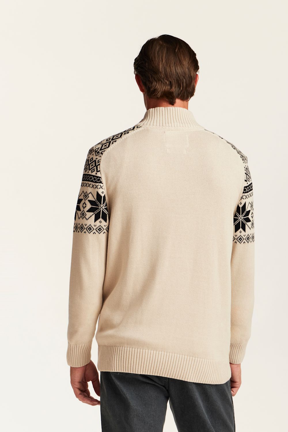 A man viewed from the back wearing a cream-colored knit sweater with black and white Fair Isle patterned yoke panels on the shoulders, paired with dark gray trousers, against a solid off-white background.