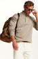 A man wearing a light beige heathered knit quarter-zip sweater, white trousers, sunglasses, and carrying a brown backpack, photographed against a clean white background.