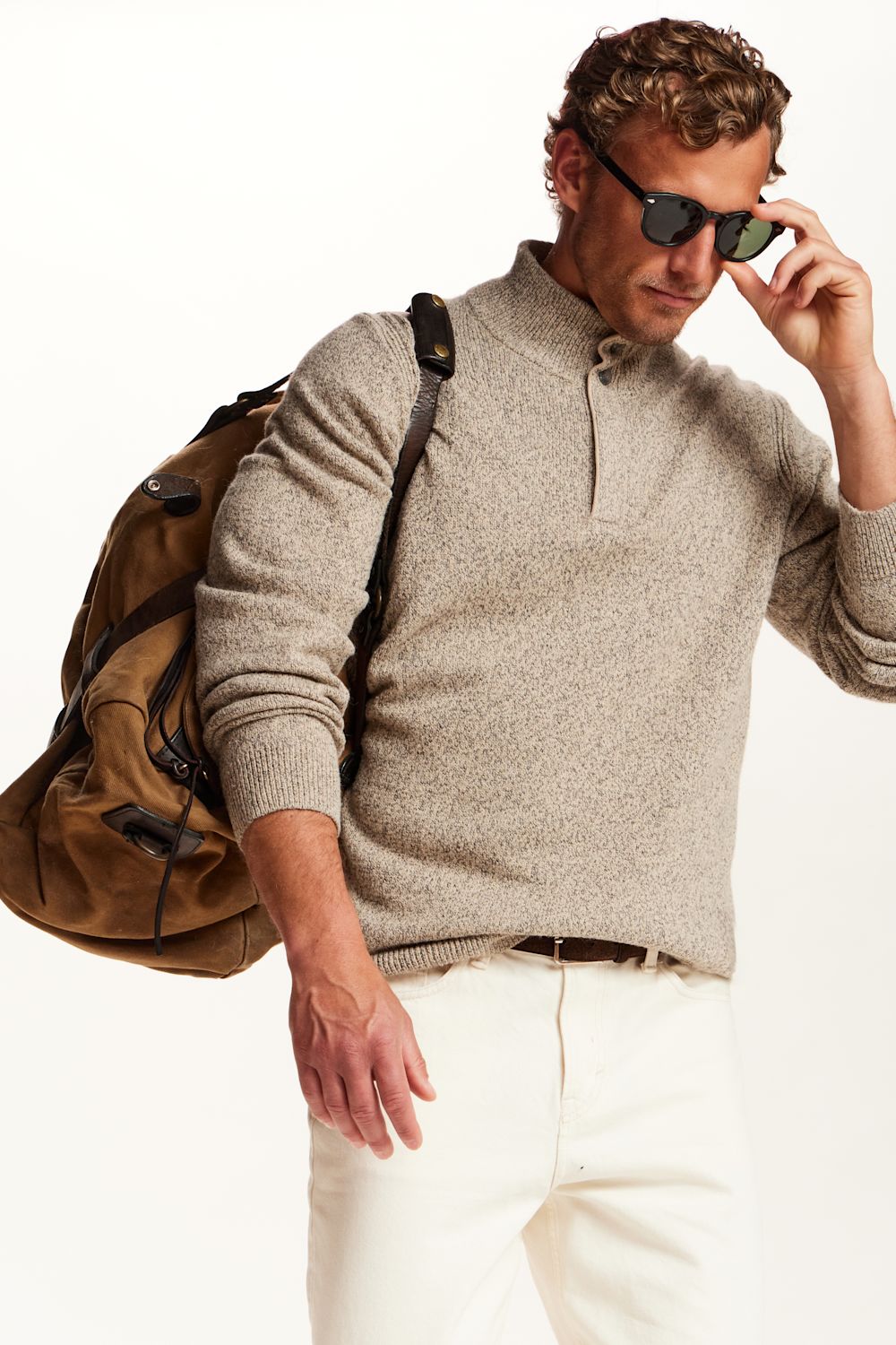 A man wearing a light beige heathered knit quarter-zip sweater, white trousers, sunglasses, and carrying a brown backpack, photographed against a clean white background.