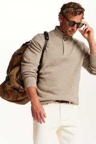 A man wearing a light beige heathered knit quarter-zip sweater, white trousers, sunglasses, and carrying a brown backpack, photographed against a clean white background.