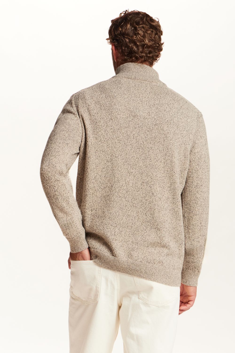 A man viewed from behind wearing a light beige textured knit sweater and off-white trousers, set against a plain white studio background.