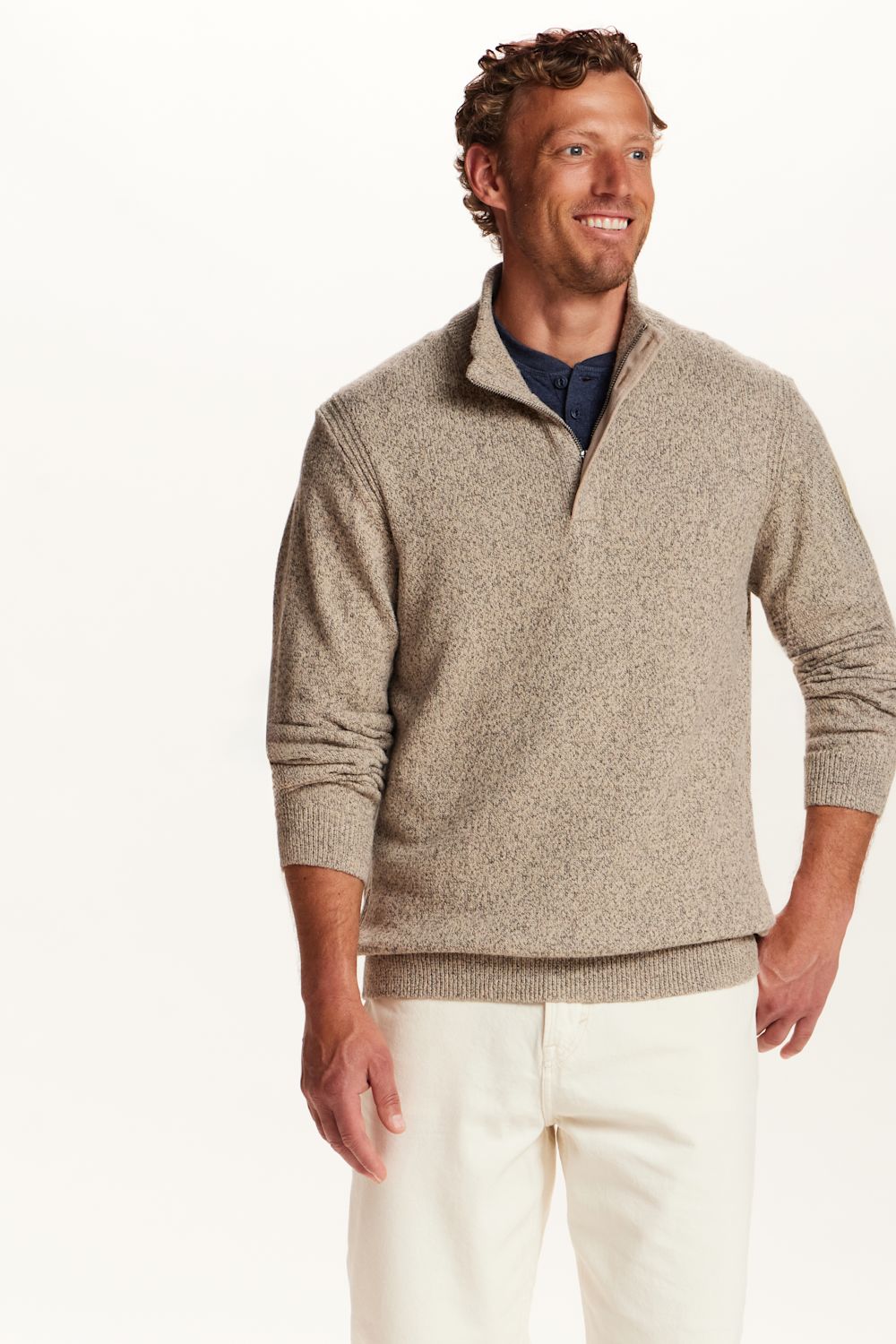 A man wearing a light beige heathered quarter-zip knit sweater over a navy blue shirt and off-white trousers, standing against a plain white studio background.