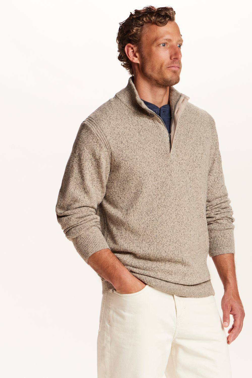 A man wearing a light beige heathered quarter-zip knit sweater over a navy collared shirt and white trousers, photographed against a clean white studio background.